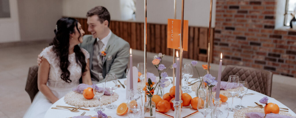 Hochzeitspaar an festlich gedecktem Tisch mit Orangen und lila Dekor