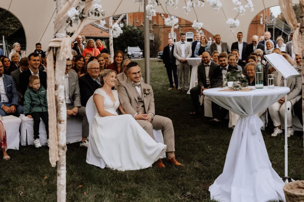 Hochzeitspaar sitzt bei Zeremonie, Gäste schauen zu